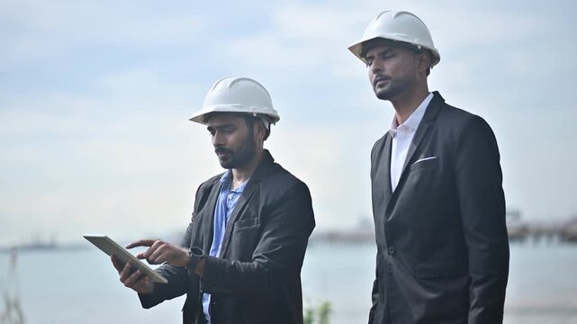 Engineers wearing safety gear, including hard hats examining survey a large blueprint tablet standing industrial facility gas or oil refinery engaged in a job requires high safety standards concept.	