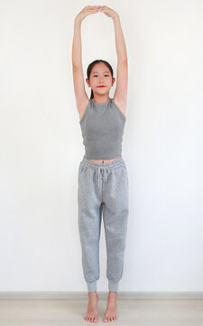Beautiful young Asian girl in comfortable grey sportswear practicing a graceful yoga pose against white wall in room.