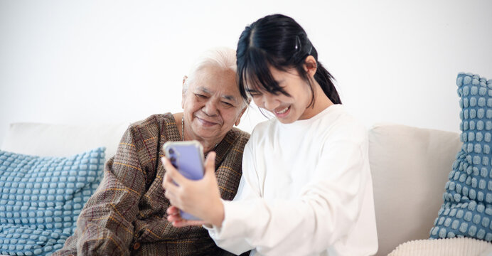Happy teenage girl showing smartphone to senior woman at home, highlighting family bonding, digital communication, technology learning, and active aging.