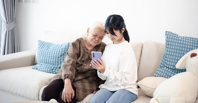 Happy teenage girl showing smartphone to senior woman at home, highlighting family bonding, digital communication, technology learning, and active aging.