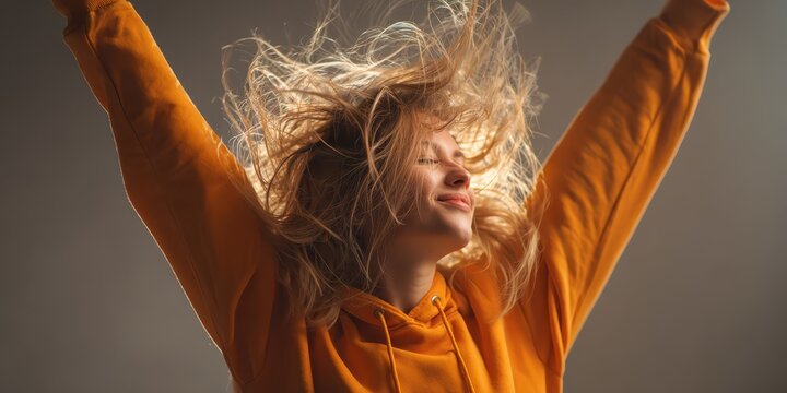 Joyful Celebration: A young woman with wind-swept hair, dressed in an orange hoodie, throws her arms up in ecstatic jubilation, her face radiant with pure happiness and freedom.