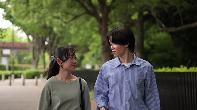話しながら新緑の歩道を歩く若いカップルのトラッキングショット