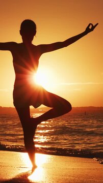 Vertical video of Yoga woman at beach sunset in tree pose meditating outdoors on by ocean sea. Female yoga instructor working out training in serene ocean landscape. Silhouette of woman doing yoga.