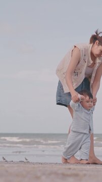 Young mother holding her baby's hands and helping him to take first steps on the sandy beach.