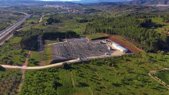 Electrical substation surrounded by green forest and agricultural land, with a highway, town and mountains visible in the background under clear blue sky