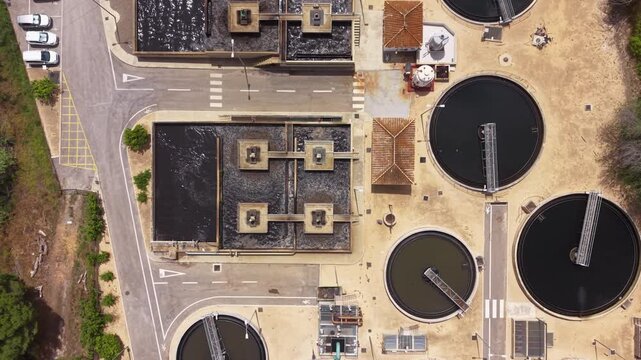 Overhead view of urban wastewater treatment plant showing circular clarifier tanks, rectangular aeration basins, access roads and facility infrastructure from above
