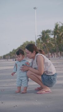 Young mother holding her baby's hands and helping him to take first steps on the sandy beach.