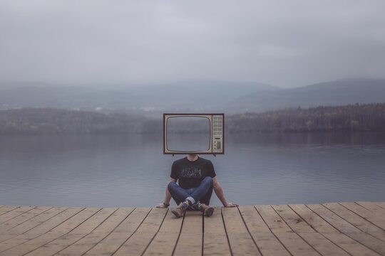 Person sitting on wooden dock with vintage tv head by water