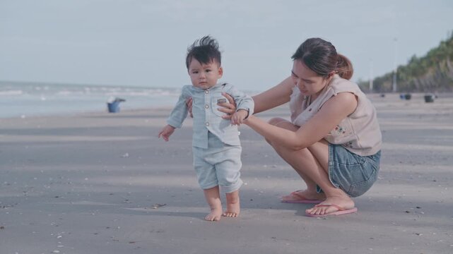 Young mother holding her baby's hands and helping him to take first steps on the sandy beach.