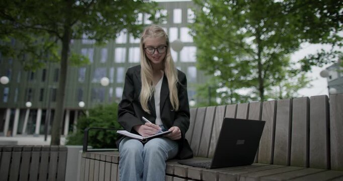 A young Caucasian female student wearing glasses in a modern urban environment among houses and trees sits on a bench in a park and works on a laptop writing notes in a notebook