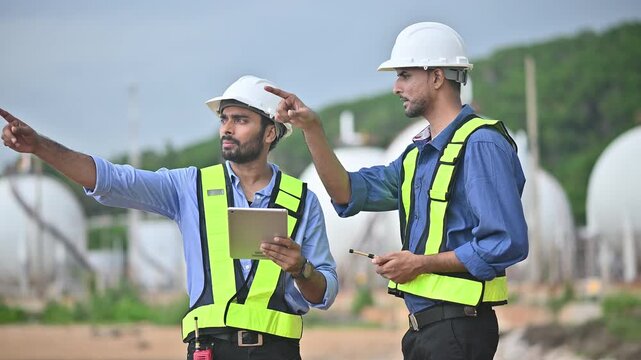 Engineers wearing safety gear, including hard hats examining survey a large blueprint tablet standing industrial facility gas or oil refinery engaged in a job requires high safety standards concept.	