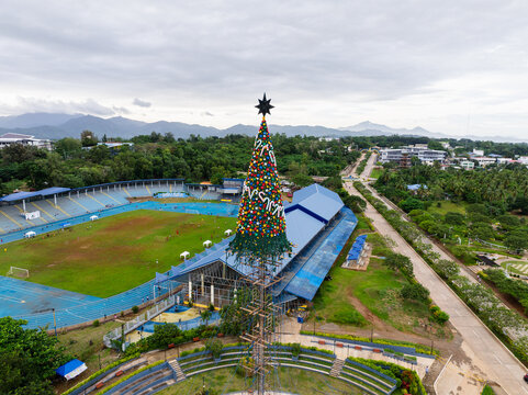 Puerto Princesa City, Philippines - 16 November 2025: Aerial view of the giant Christmas tree at Balayong Rotonda Park with the city sports complex and distant mountains under a cloudy sky.