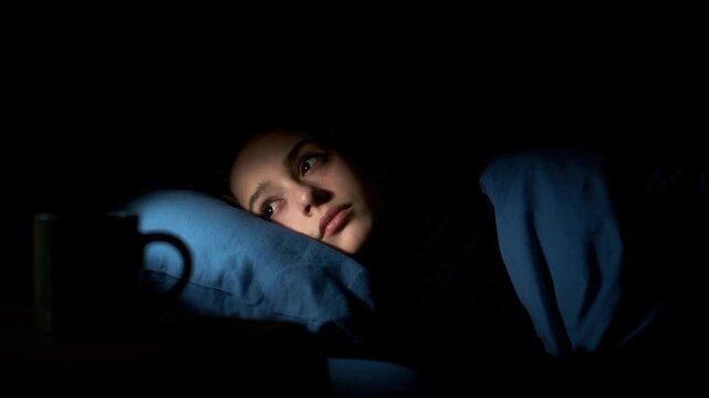 Young woman lying awake in bed at night with worried expression, struggling with insomnia and inability to sleep in dark bedroom environment