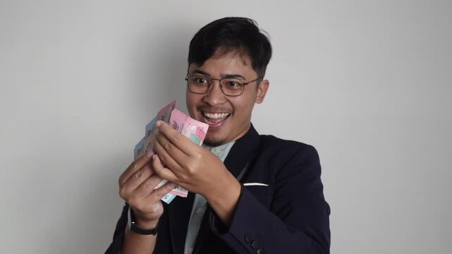 A young man in a black blazer and glasses looks excitedly at a fan of Indonesian rupiah banknotes, symbolizing financial success, profit, and a positive outlook on money.