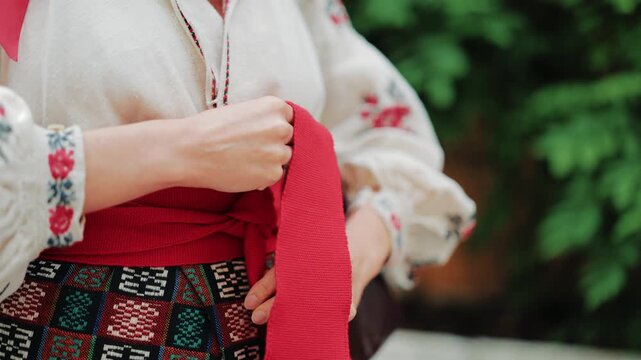 Detailed close-up of a woman's hands tying a vibrant red sash over an authentic Ukrainian vyshyvanka. Showcasing traditional folk clothing details, ethnic craftsmanship, and heritage fashion design.