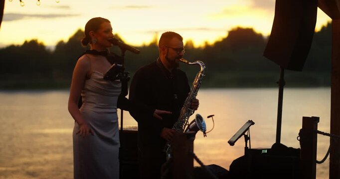 Duet performance by female singer and male saxophonist at lakeside sunset