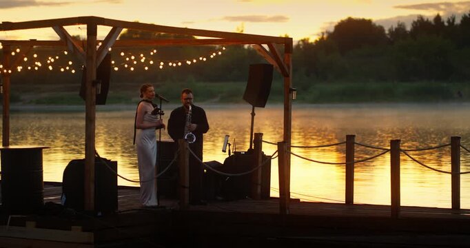 Sunset Duo Performing on Wooden Lakeside Pier Under String Lights