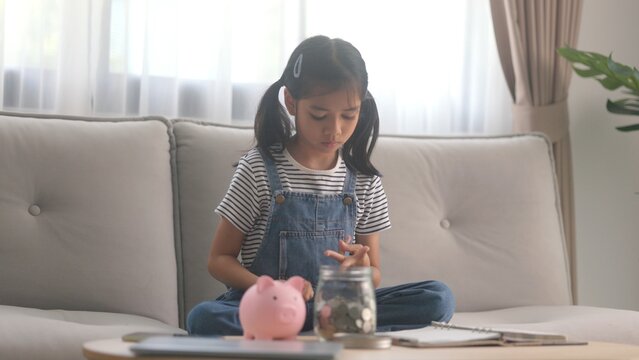 Smart female child counting saved coins next to glass jar on table. Cute little girl practicing basic economics and budget deposit planning inside cozy living room. Essential lifestyle content.