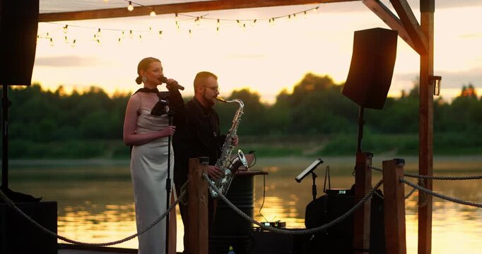Female Singer and Saxophonist Performing on Lakeside Pergola at Sunset