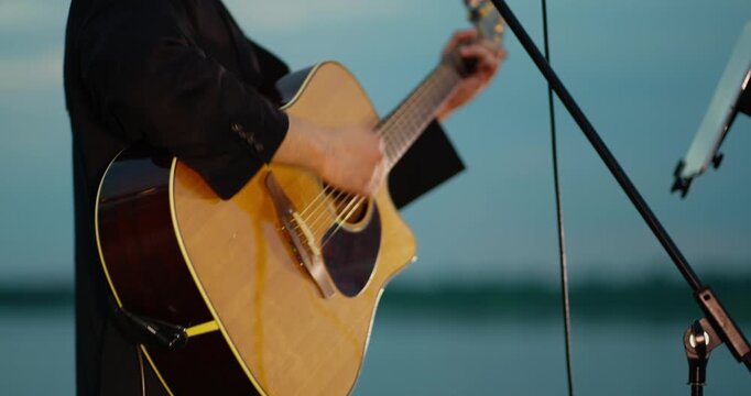 Close-up of musician strumming acoustic guitar on outdoor stage at dusk