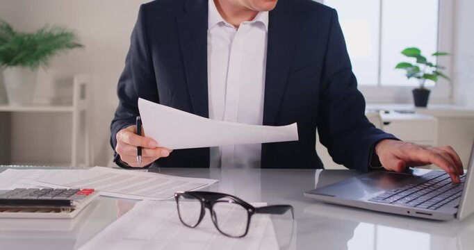 Portrait of business man accountant wearing suit with glasses on table working with financial documents on his workplace looking at the laptop monitor screen sitting in office. 4k video.