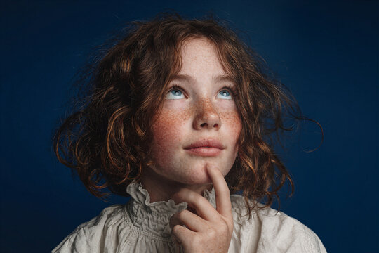 Young girl with freckles thoughtfully looking up with finger on chin