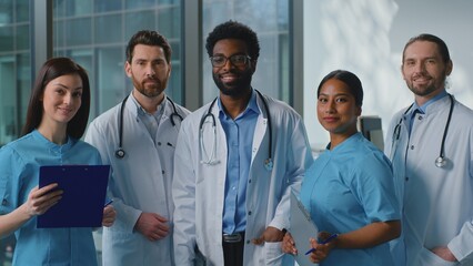 Fototapeta premium Happy smiling multiethnic doctors look in digital tablet talking discussing online health test results in clinic. Joyful team five surgeons looking at camera smile talk medicine teamwork in hospital