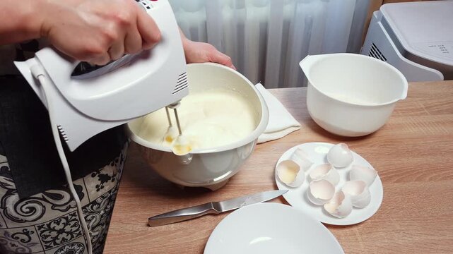 A person uses an electric hand mixer to blend ingredients in a white bowl, with eggshells nearby, suggesting baking or cooking