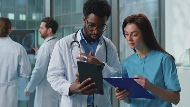 Two diversity doctors man with woman discussing medical examination using digital tablet together in hospital. Caucasian female therapist doctor talking with African American male surgeon at clinic