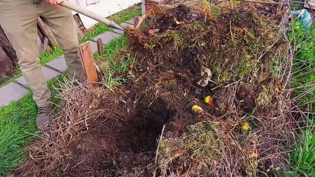A person uses a pitchfork to aerate and mix organic materials in a garden compost bin, promoting decomposition for nutrient-rich soil