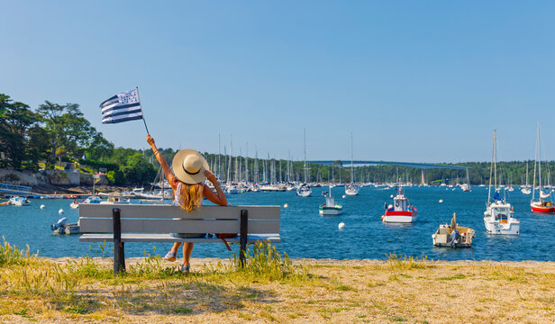 Back view of female tourist sitting on bench waving brittany flag looking at boats in regional harbor