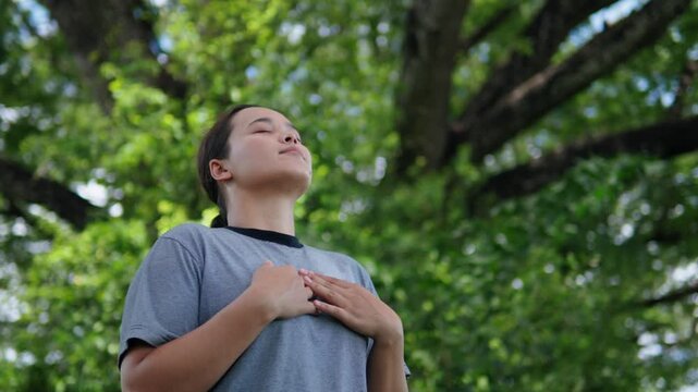 An Asian woman in a striped shirt closes her eyes and takes a deep breath to relax with the sounds of nature, healing her heart surrounded by lush greenery.