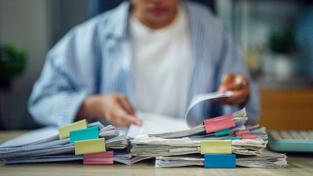 Person sorts through stacks of papers marked with colorful sticky notes. Focus is on hands and documents, not face or surroundings. Workload suggests busy office or administrative tasks