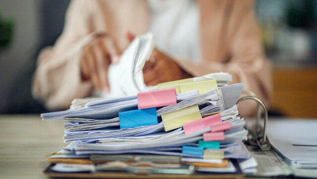 Person sits at desk, overwhelmed by paperwork. Hands clutch phone, eyes focused on screen. Stack of files topped with colorful sticky notes. Busy office environment, cluttered with documents