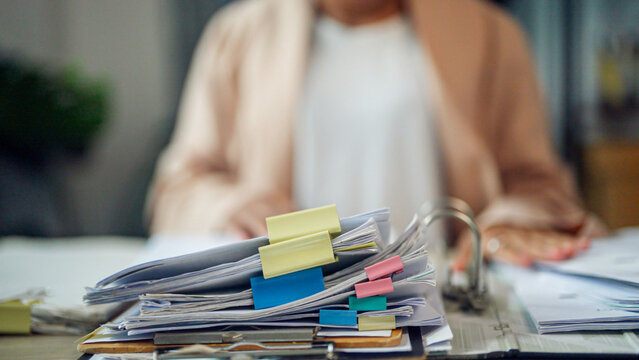 Blurry person sits at desk, hands resting nearby. Stack of papers, colorful clips, and binders dominate foreground. Office clutter suggests busy workday or pending tasks