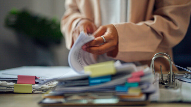 Hands flip through papers with focused intent. Stacks of documents, sticky notes, and folders lie nearby. Soft lighting highlights organized chaos at a desk. Subtle ring glints on a finger