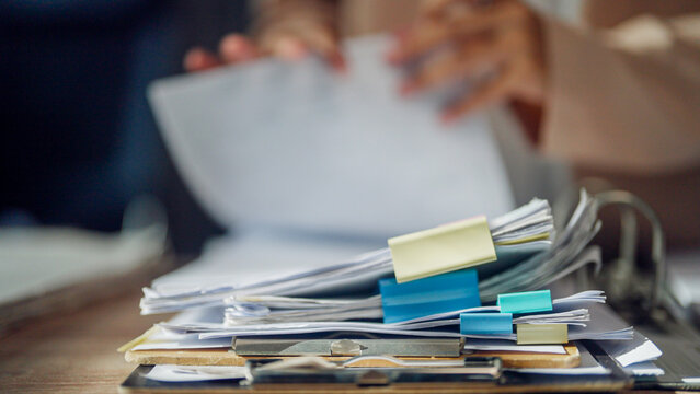 Hands hold papers above a cluttered desk. Piles of files, clips, and folders fill the surface. The scene suggests busy work or overwhelmed tasks. Soft focus hints at stress or urgency