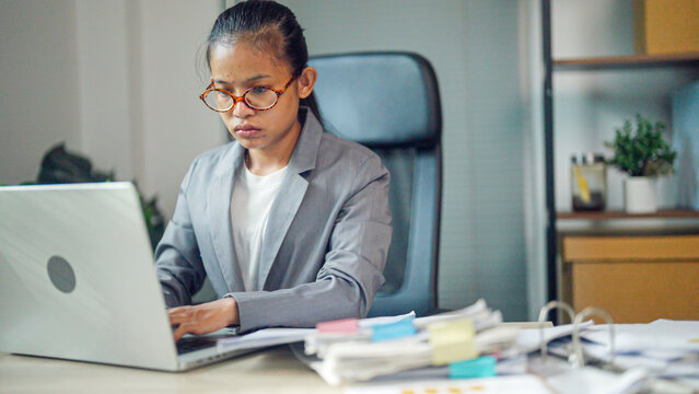 Focused woman types on laptop at cluttered desk. She wears glasses and a tailored blazer. Soft light illuminates her concentrated expression. Documents and sticky notes surround her workspace
