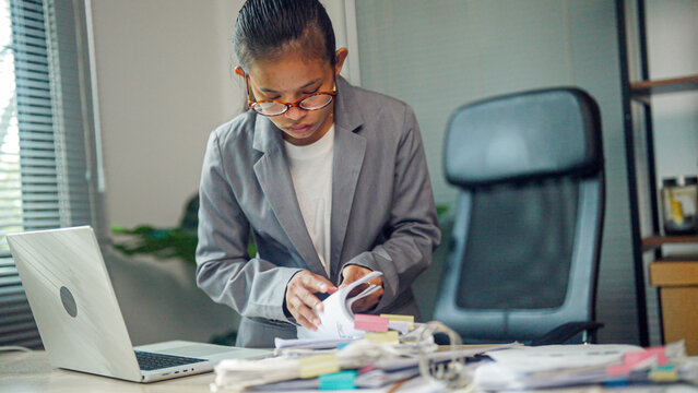 Young professional organizes paperwork in tidy office space. Focused expression suggests dedication to task. Laptop and desk clutter imply busy workload. Ideal for business, management