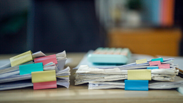 Stacks of papers, colorful tabs, and a calculator sit on a desk. The scene suggests busy work or urgent organization. Focus is on details, not people or emotions. Ideal for business, finance