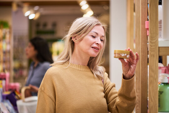 Caucasian middle aged woman examining cosmetic product on beauty store shelf while shopping. Suitable for retail marketing, skincare promotion, consumer choice concept