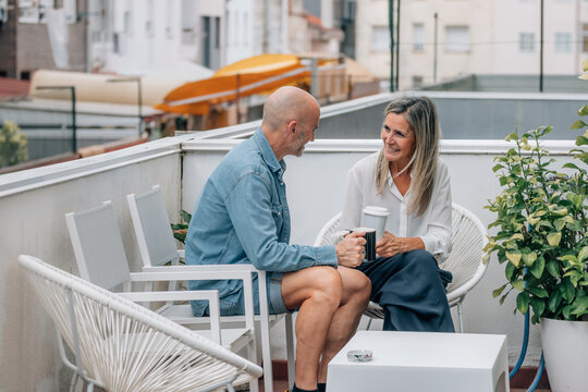 Two caucasian middle-aged adults share a joyful moment over coffee on a sunlit outdoor terrace, radiating warmth, connection, and relaxed lifestyle