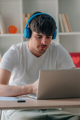 young man at home taking an online course with headphones and a laptop on his desk, studying and...