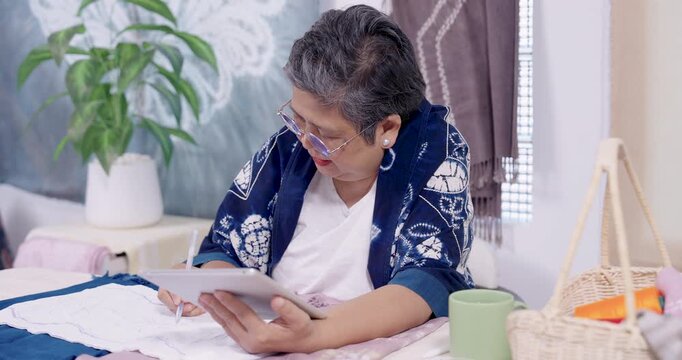 Senior Asian craft entrepreneur leans low, studies tablet, then writes pattern mark on fabric, refining shibori resist stitching guide for indigo dye process, sustainable home studio order work.