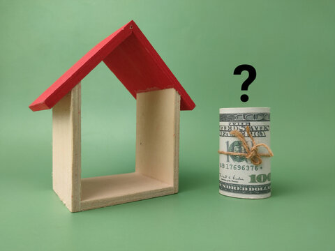 A miniature wooden house frame stands next to a rolled dollar bundle beneath an overlaid black question mark