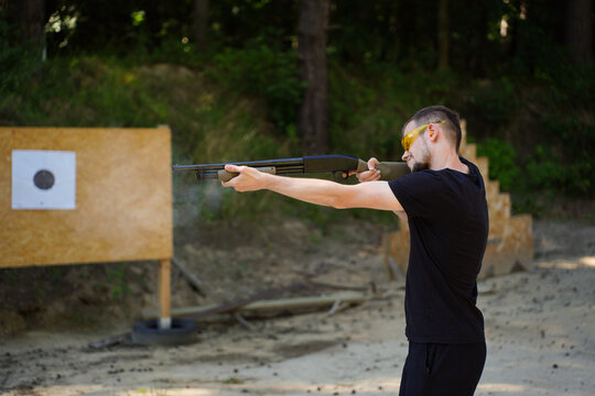 Man in black t-shirt shooting a pump-action shotgun at a target range in an outdoor forest setting. Firearm marksmanship training, shotgun sport practice, outdoor shooting range discipline 