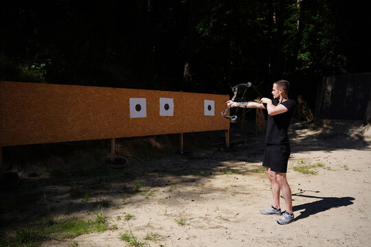 Young man practicing archery aiming compound bow at targets on wooden backstop in outdoor field. Archery training, bowhunter marksmanship practice, outdoor shooting range sport 