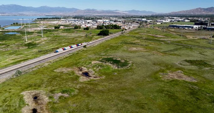 The Frontrunner light rail commuter train gliding past fields near Vineyard, Utah, with green grass, Utah Lake, and the Wasatch Front on it's way from Provo to Ogden - aerial flyover