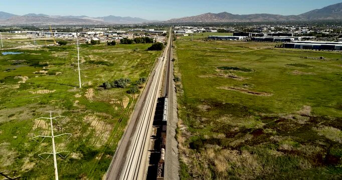 Forward drone footage follows a Union Pacific cargo train traveling across green fields near Vineyard, Utah towards the Salt Lake City valley and Wasatch Mountains ion a sunny day