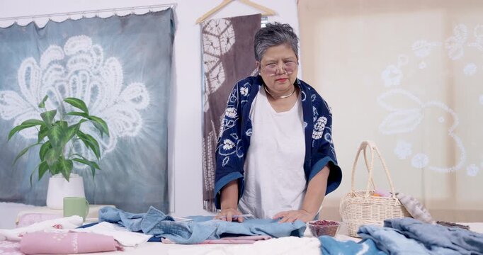 Senior Asian woman artisan stands with tablet, sorts denim fabric pieces on table, checks inventory for small business order, prepares shibori resist stitching session before indigo dyeing step.